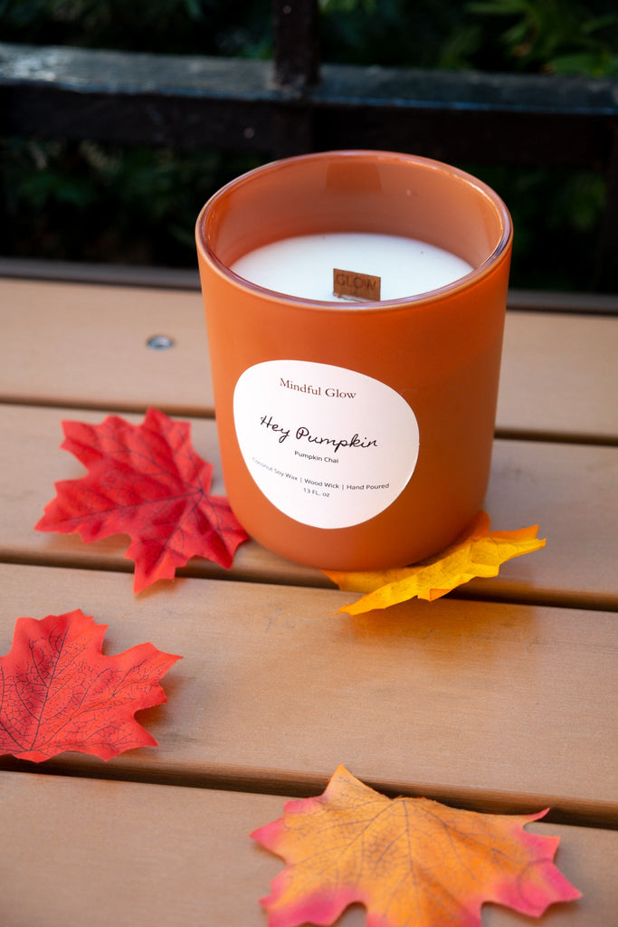 The Mindful Glow - Hey Pumpkin Woodwick Candle 13oz sits on a wooden surface with faux autumn leaves, adding cozy fall vibes. Made by Mindful Glow with coconut soy wax, the candle is perfect for autumn décor. Greenery is softly blurred in the background.