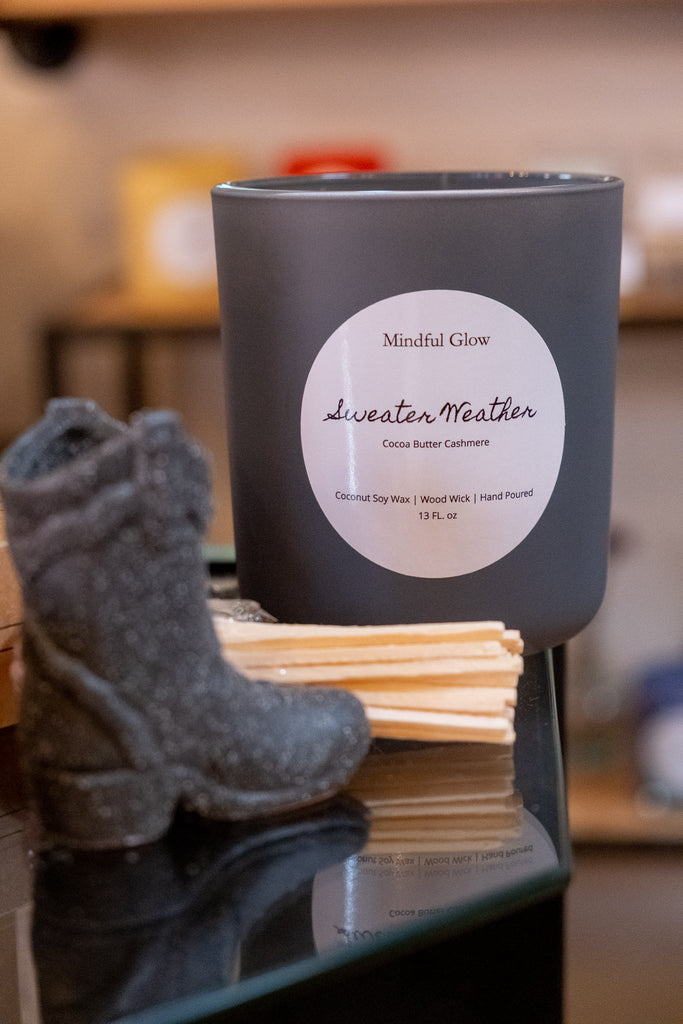 A 13oz Mindful Glow - Sweater Weather Candle with a cozy cocoa butter cashmere scent rests on glass beside a black cowboy boot and wooden sticks, ideal for relaxing aromatherapy.