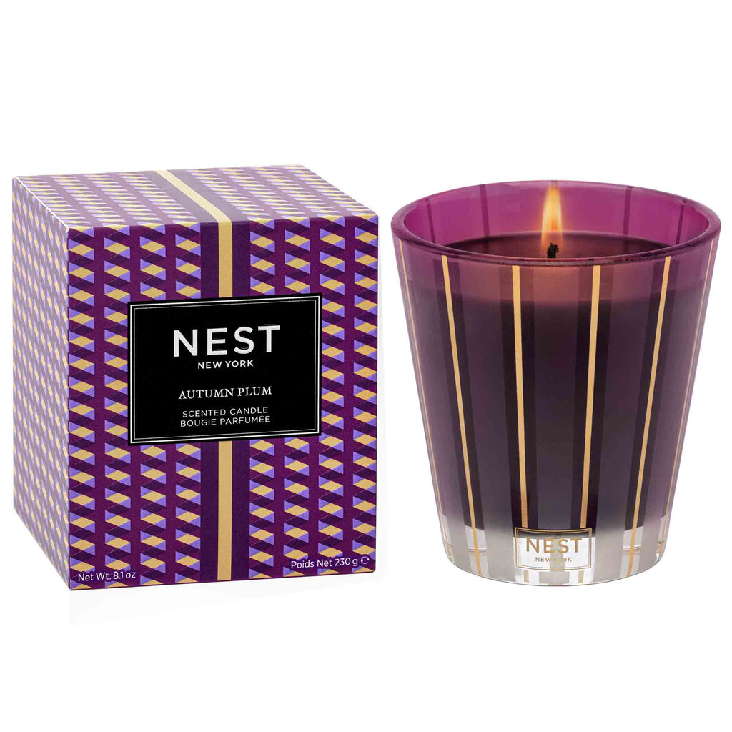 The lit Nest - Autumn Plum Classic Candle from Nest emits a luxurious, fruity wood scent. This premium purple glass candle, featuring gold stripes, sits beside its matching geometric box labeled “NEST New York Autumn Plum Scented Candle.”.