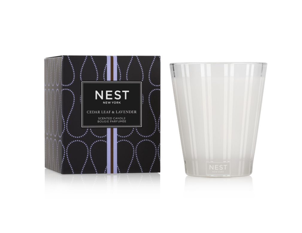 The Nest - Cedar Leaf & Lavender Classic Candle, in white glass, is shown next to its black box adorned with a purple dotted pattern and a white label displaying the scent name.