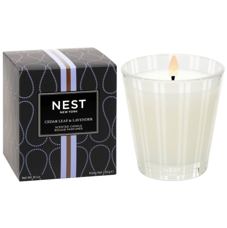 A lit Nest - Cedar Leaf & Lavender Classic Candle in a clear glass holder sits beside its black box, which is decorated with silver lines and dots. The packaging displays the Nest brand name.