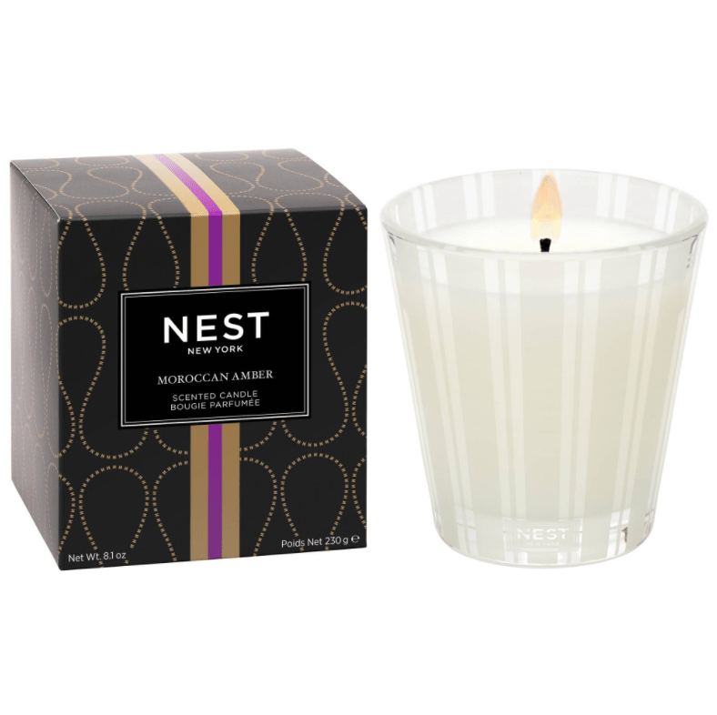 The Nest - Moroccan Amber Classic Candle by Nest burns in a clear glass jar beside its black and gold box with a purple stripe, filling the room with a rich, woody fragrance.