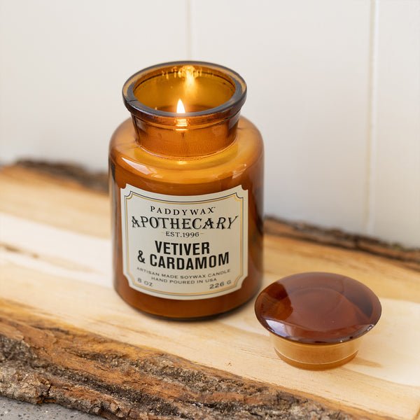 A Paddywax - Apothecary - Vetiver + Cardamom 8oz Candle burns on a wooden surface, its amber glass jar and lid reflecting vintage apothecary style—a timeless home fragrance by Paddywax.