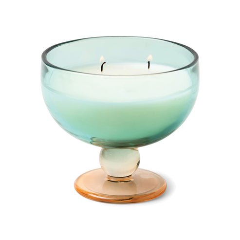 The Paddywax Aura Candle in Tobacco Patchouli, with its light blue vintage glassware and amber pedestal base, holds a lit white two-wick candle. It emits a warm glow while hints of spices fill the air, creating a cozy ambiance.