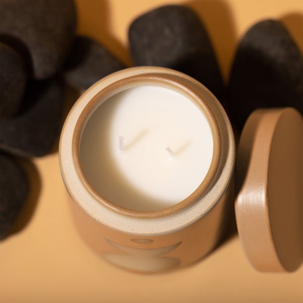 The Paddywax - Form - Wild Fig + Vetiver 12oz Candle, featuring two wicks, sits open beside its lid and is surrounded by smooth black stones on a beige surface.
