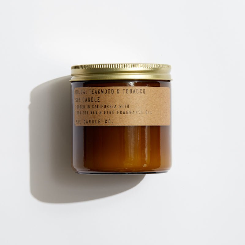 The P.F. Candle Co. - 12.5 oz. Teakwood and Tobacco Candle, housed in a brown glass jar with a gold lid, offers a woody aroma. The label highlights its teak-infused California production as it rests against a white background, ready to fill your space with its rich scent.