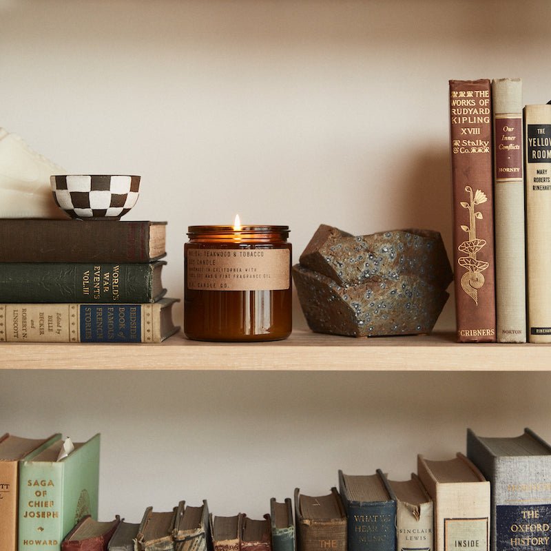 Next to a rock and checkered bowl, a P.F. Candle Co. - 12.5 oz. Teakwood and Tobacco Candle emits a woody scent by a cozy bookshelf lined with colorful books, including works by Rudyard Kipling, on the teak shelf.