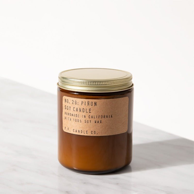 A brown glass jar with a gold lid holds the P.F. Candle Co. - 7.2 oz. Piñon Candle, handmade in California from 100% soy wax and fine fragrance oils, offering a warm, woody scent.