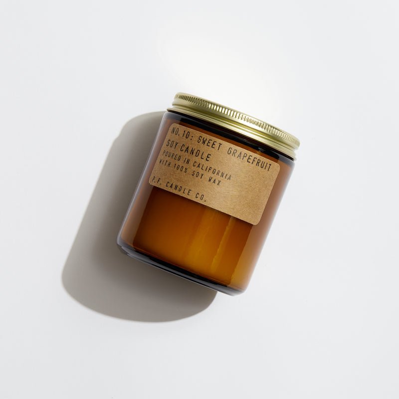 The P.F. Candle Co. Sweet Grapefruit Candle is a 7.2 oz soy wax candle in a brown glass jar with a gold lid, hand-poured in California and paraben-free. The jar, casting a shadow on light surfaces, exudes elegance and simplicity with its fruity scent.