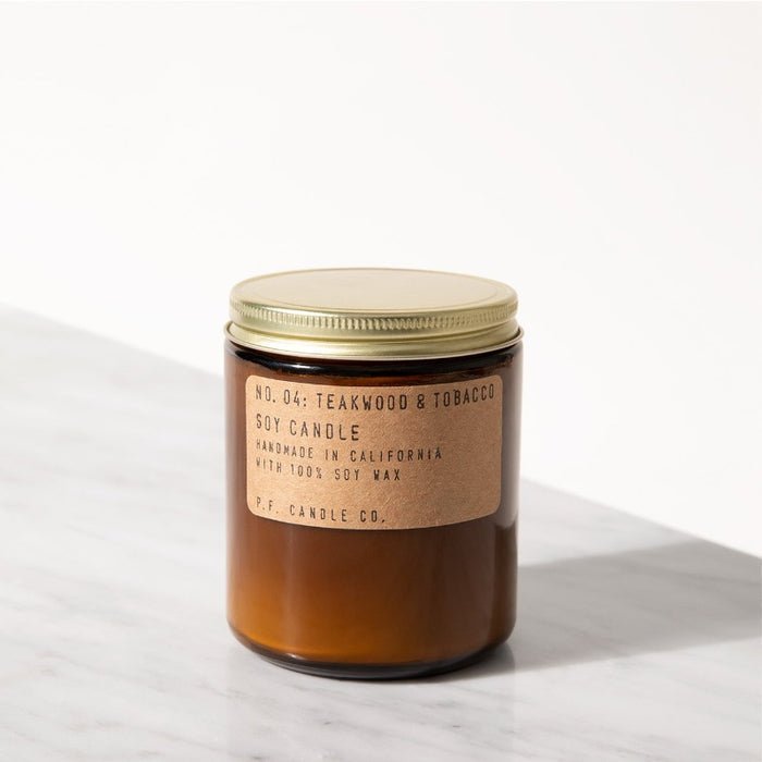 A P.F. Candle Co. 7.2 oz. Teakwood and Tobacco candle in a brown glass jar with gold lid rests on white marble, filling the air with leather and woody notes. The label highlights its handmade in California from 100% soy wax.