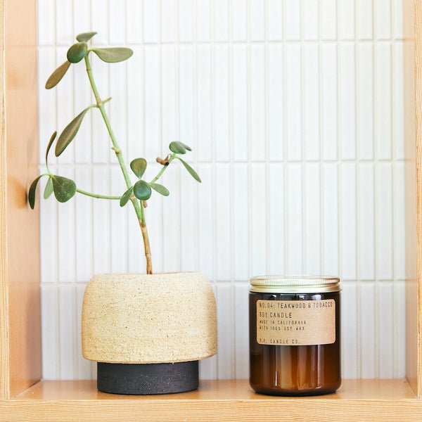 A small potted plant sits beside a P.F. Candle Co. - 7.2 oz. Teakwood and Tobacco Candle, featuring a woody soy wax scent, on a wooden shelf against white tiles.