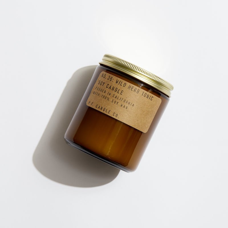 A brown glass jar candle with a gold lid labeled Wild Herb Tonic, No. 36 by P.F. Candle Co., crafted from soy wax in California and infused with rich fragrance oils, casts a soft shadow on the light background.