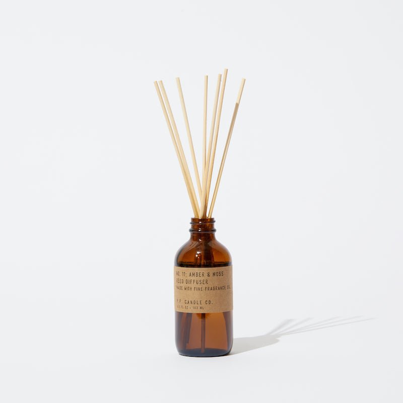 The P.F. Candle Co. - Diffuser - Amber & Moss features a brown glass bottle with a kraft label, filled with natural reed sticks that diffuse subtle amber and moss scents, displayed against a white background.