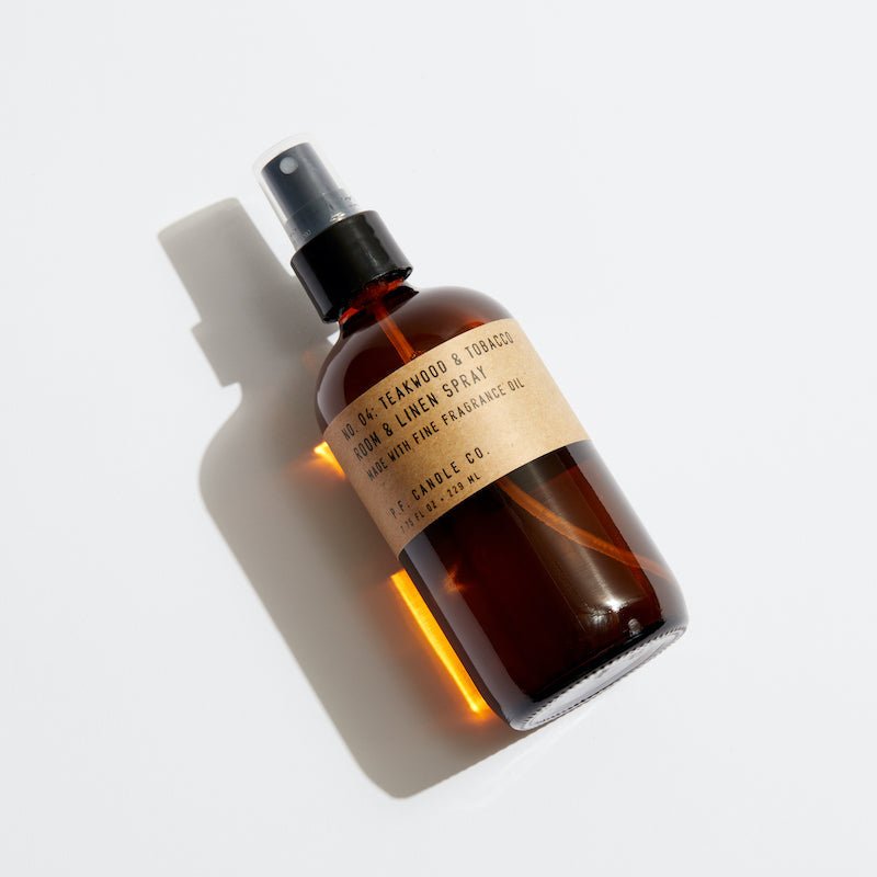 A brown glass spray bottle with a black nozzle, labeled P.F. Candle Co. - Linen & Room Spray - Teakwood & Tobacco, releases a woody fragrance. It casts a shadow on a white background, suggesting subtle notes of leather and teak entwined.