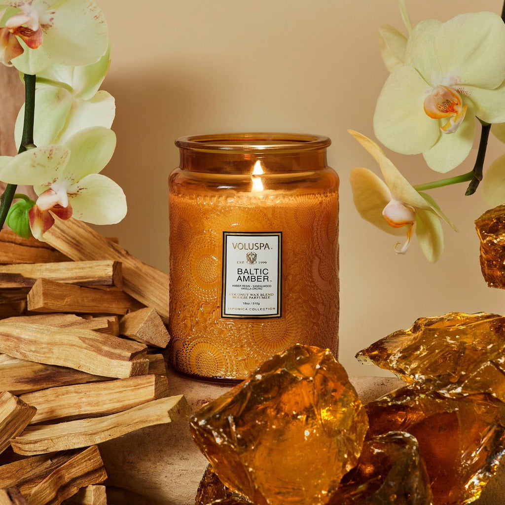 A Voluspa Baltic Amber Large Jar 18oz Candle, crafted with a coconut wax blend, rests on a beige background among light wooden pieces, yellow orchids, and amber-colored stones. Housed in an ornate amber glass jar with a white label, it exudes comforting notes of vanilla orchid.