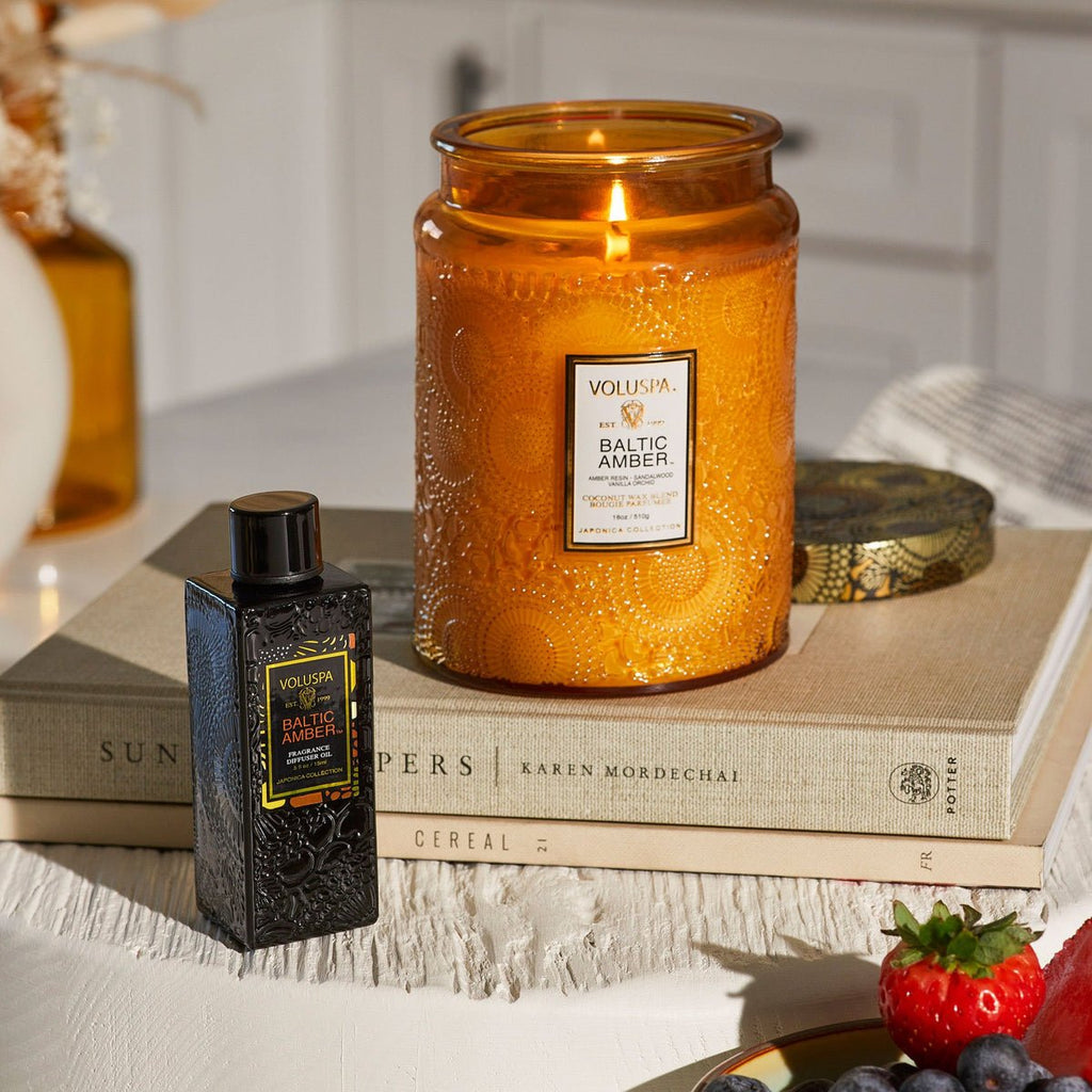 A lit Voluspa Baltic Amber Large Jar 18oz Candle sits atop closed books. Beside it, a matching ornate bottle adds elegance. Strawberries and grapes lie on a sunlit white table, as the vanilla orchid and amber resin scent completes the tranquil atmosphere.