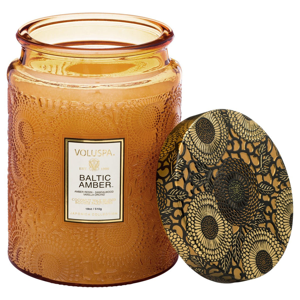 An 18oz Voluspa Baltic Amber glass jar candle, amber-colored and lidless, sits beside its black and gold floral-patterned lid. Crafted from a coconut wax blend, the jars textured design adds elegance.