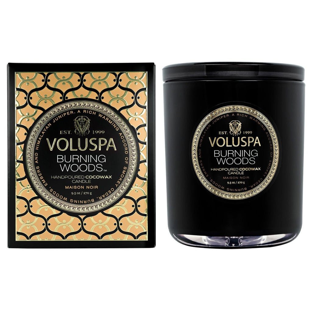 The Voluspa Burning Woods Classic Candle 9.5oz comes in a black jar within an ornate black and gold box, labeled Handpoured Vegan Coconut Wax Candle, featuring details on size and fragrance. Its elegant design captures the essence of smokey cedar.