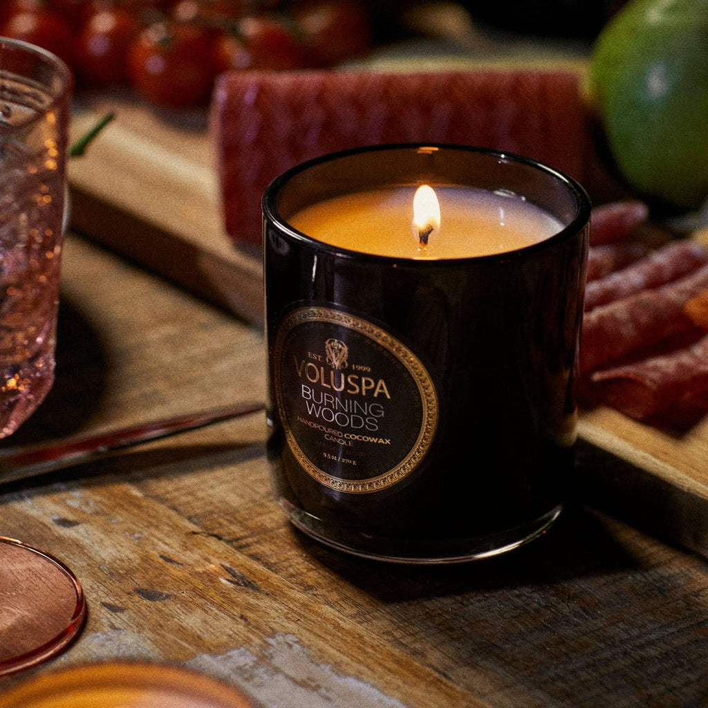 A Voluspa - Burning Woods Classic Candle 9.5oz with coconut wax glows warmly on a wooden table amidst cherry tomatoes and cured meats, infusing the air with a hint of smokey cedar and creating a cozy atmosphere.