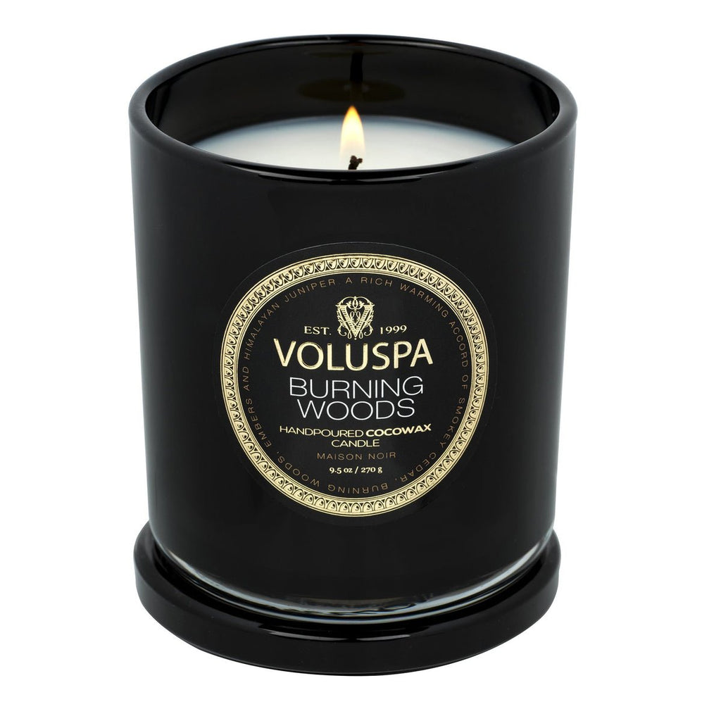 A Voluspa - Burning Woods Classic Candle in glossy black glass emits a smokey cedar aroma. Featuring Handpoured Coconut Wax and Maison Noir on a gold and black label, this sophisticated 9.5oz vegan candle offers elegance with its lit wick.