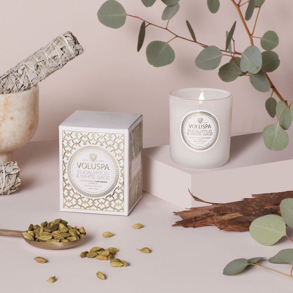 A lit Voluspa Eucalyptus and White Sage Classic Candle (9.5oz) made from coconut wax rests beside its decorative box, surrounded by sage, cardamom pods, and eucalyptus leaves on a neutral surface.