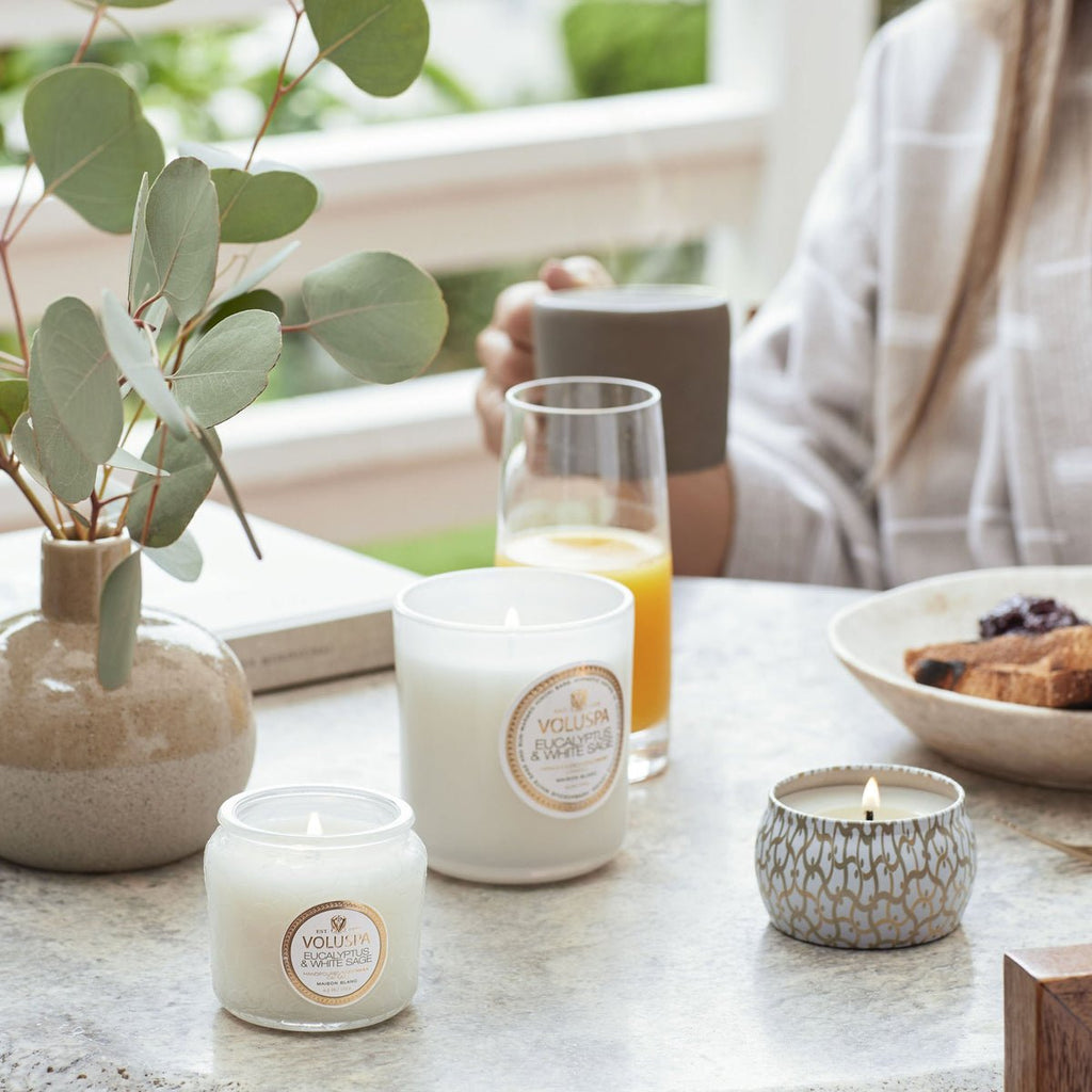 A table with three lit Voluspa - Eucalyptus and White Sage Classic Candles in jars, a small vase of eucalyptus branches, a glass of orange juice, and pastries. A person holds a partially visible mug while natural light filters through the nearby window.