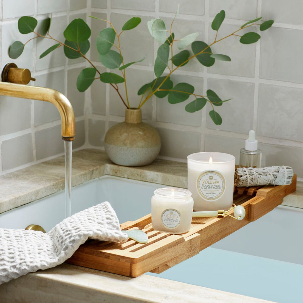 A bathtub with running water features a bamboo tray holding a Voluspa - Eucalyptus and White Sage Classic Candle, a rolled towel, bath oil, and a gold spoon. Nearby, eucalyptus branches in a ceramic vase adorn the tub ledge against beige tile walls.