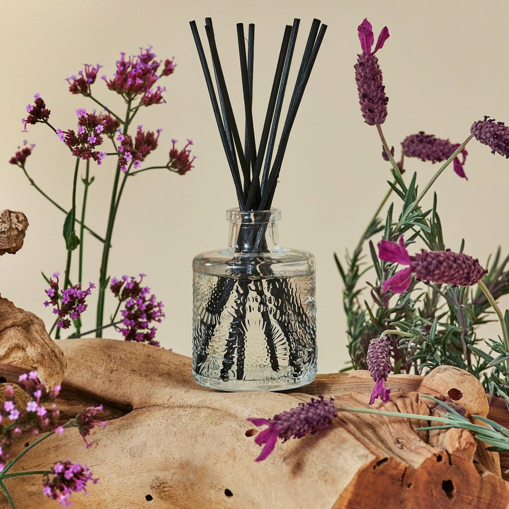 The Voluspa - French Cade Lavender 3.4oz Reed Diffuser features black sticks in a glass holder on driftwood, surrounded by purple Verbena and lavender, with a beige background enhancing its natural, rustic look and the soothing aroma of French Cade Wood.