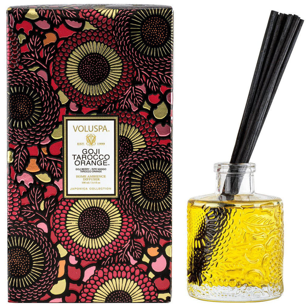 A Voluspa - Goji Torocco Orange Reed Diffuser set features vibrant floral packaging in red, black, and gold. The clear glass bottle holds yellow liquid with a tangy essence of ripe mango and includes several black reeds.