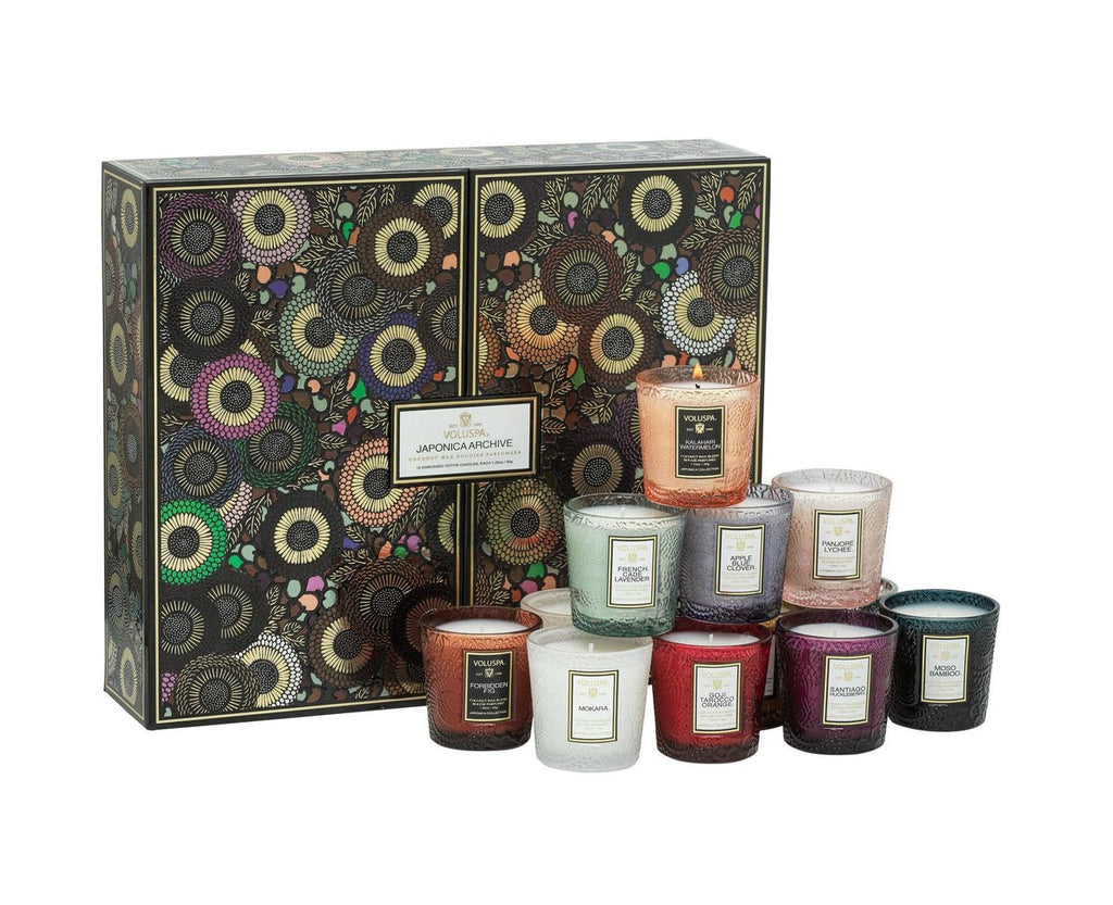 The Voluspa Japonica 12 Votive Candle Gift Set features a decorative box filled with luxury candles made from a coconut wax blend. Each colorful glass candle comes with scents like Goji Tarocco Orange and French Cade Lavender, beautifully displayed in front of the box.