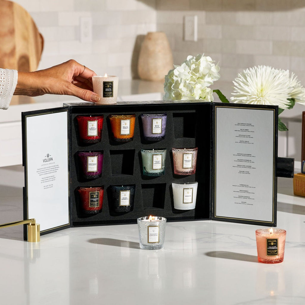 A person holds a lit Voluspa candle from the Japonica 12 Votive Candle Gift Set in front of nine assorted, colorful candles in a sleek black case. Made from coconut wax blend, they are neatly arranged with white flowers on a white kitchen counter.