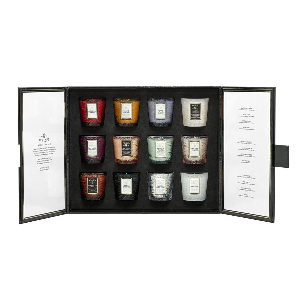 The Voluspa - Japonica 12 Votive Candle Gift Set reveals a luxurious black box with 12 vibrant coconut wax blend candles. Each candle is in an elegant glass holder. The sides of the box feature descriptions and small hinges, making it the perfect luxury gift.