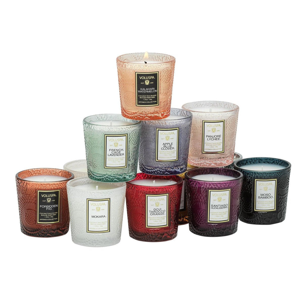 A 12-candle gift set from the Voluspa Japonica Collection is artistically arranged in a pyramid, featuring coconut wax blend candles in textured glass jars of vibrant colors like red, blue, green, and clear. With one lit at the top, they deliver an exquisite fragrance throw.