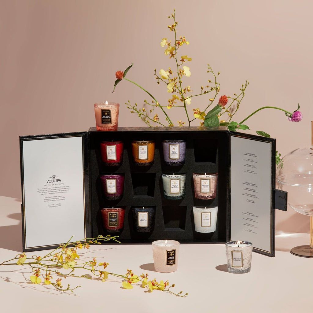 A Voluspa Japonica 12 Votive Candle Gift Set in a black box is open on the table. Two coconut wax blend candles are outside the luxe gift box, with one lit. Yellow and pink flowers in a vase and scattered blooms enhance the elegant scene.