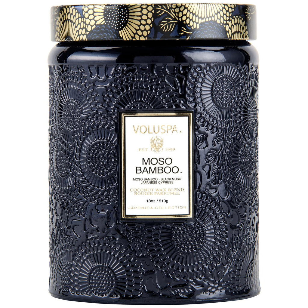The Voluspa Moso Bamboo Large Jar Candle, part of the Japonica Collection, features a decorative black jar with an embossed floral design, a golden lid, and holds 18oz. It is infused with Japanese Cypress essence in a coconut wax blend.